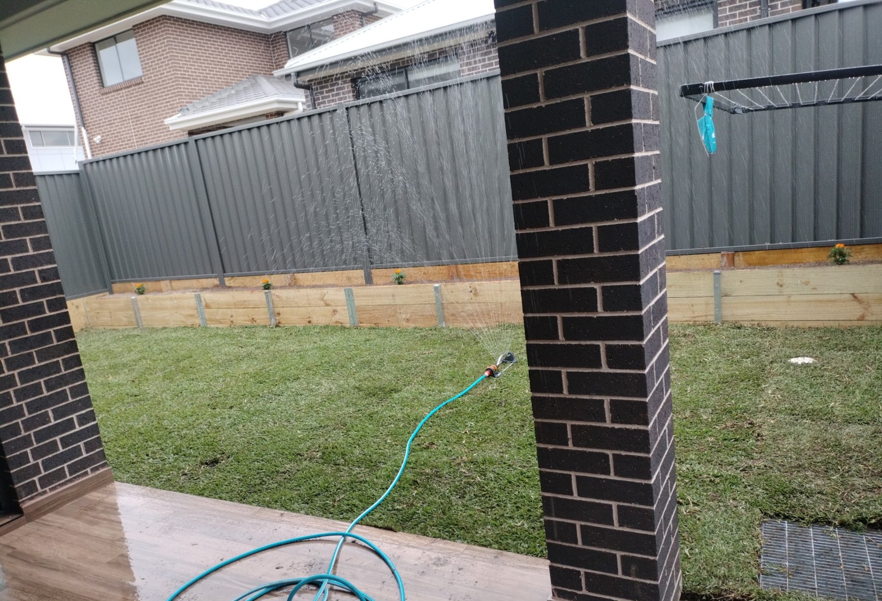 Back yard seen between two brick pillars: new turf being watered, a timber sleeper edge and grey fence